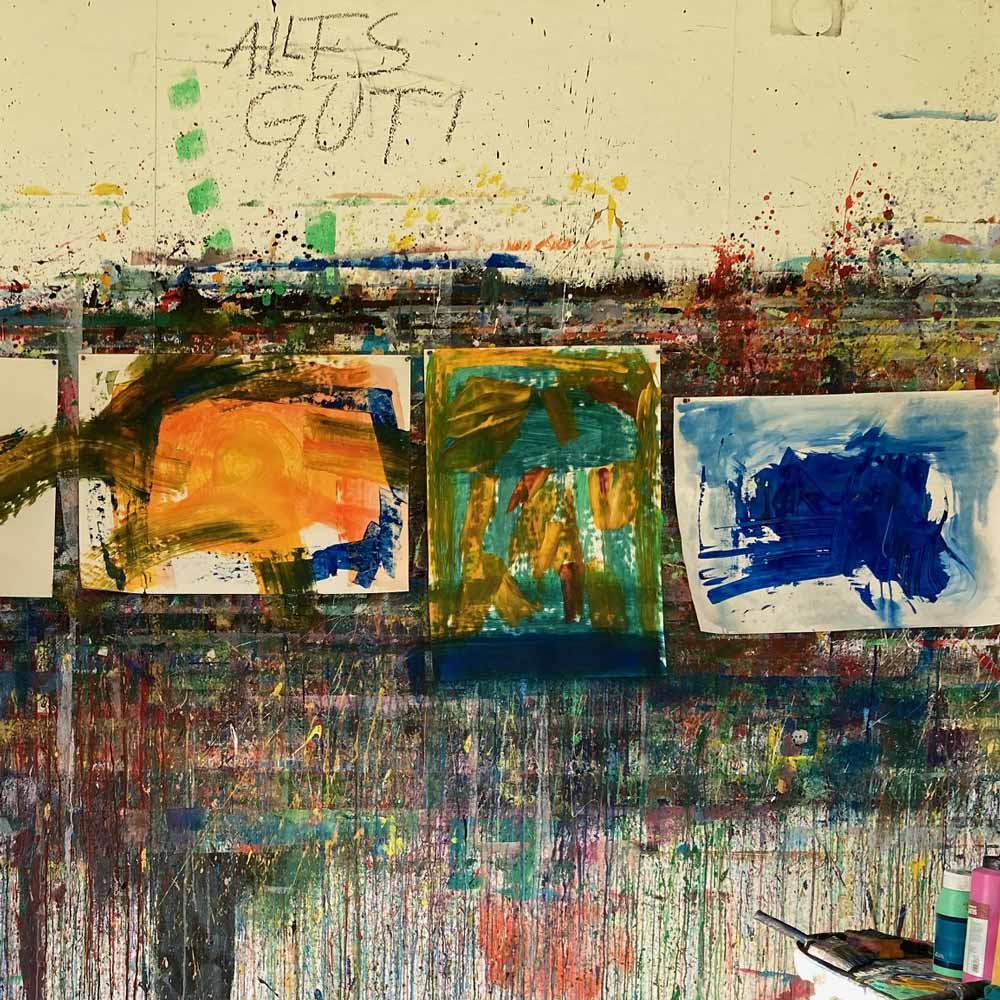 Eine Wand mit mehreren abstrakten Kunstwerken, die in intensiven Farben wie Orange, Blau und Grün gemalt sind. Die Wand ist bunt und mit Farbspritzern bedeckt, darüber steht der Schriftzug 'Alles gut!' in grüner Kreide.