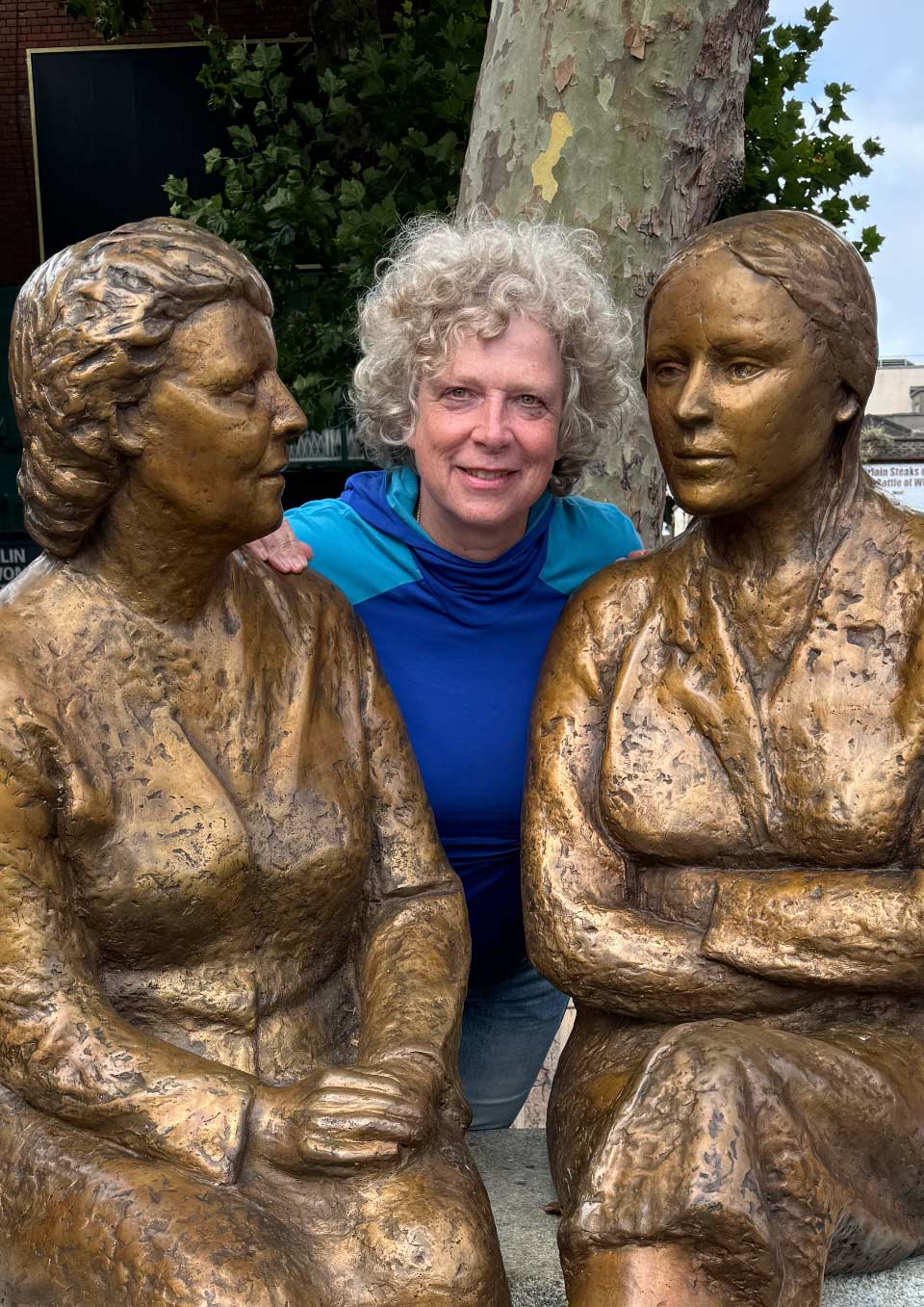 Susanne (Frau mit grauem, lockigem Haar) posiert zwischen zwei Bronzestatuen, die Frauen in sitzender Haltung darstellen.
